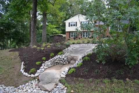 A stone-lined dry creek bed leads toward a two-story suburban house surrounded by trees and newly mulched landscaping.