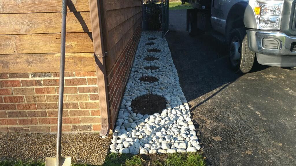 A narrow landscaping strip with white decorative rocks and five circular dirt patches, situated between a wall and driveway.