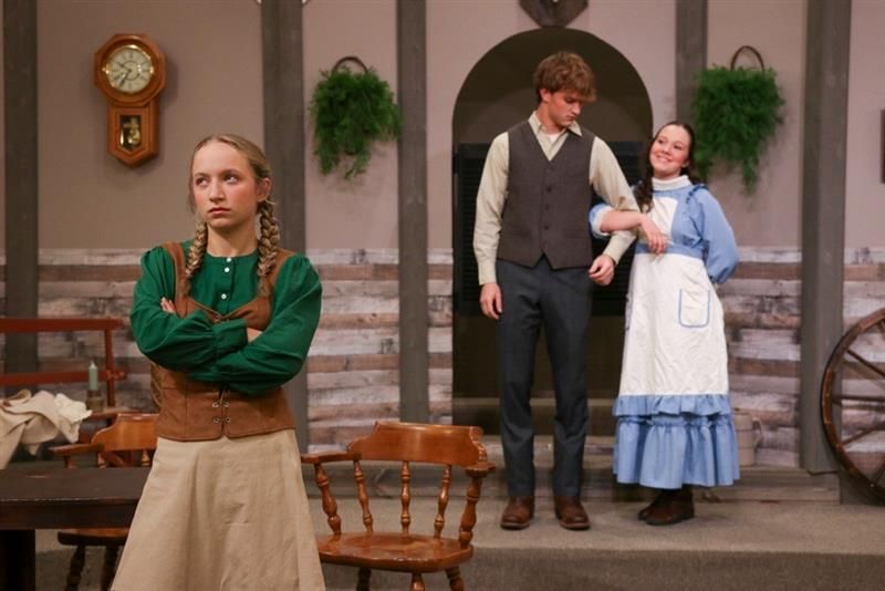 Two actors in period costume stand on a stage, with a woman in the foreground and a couple near the back.
