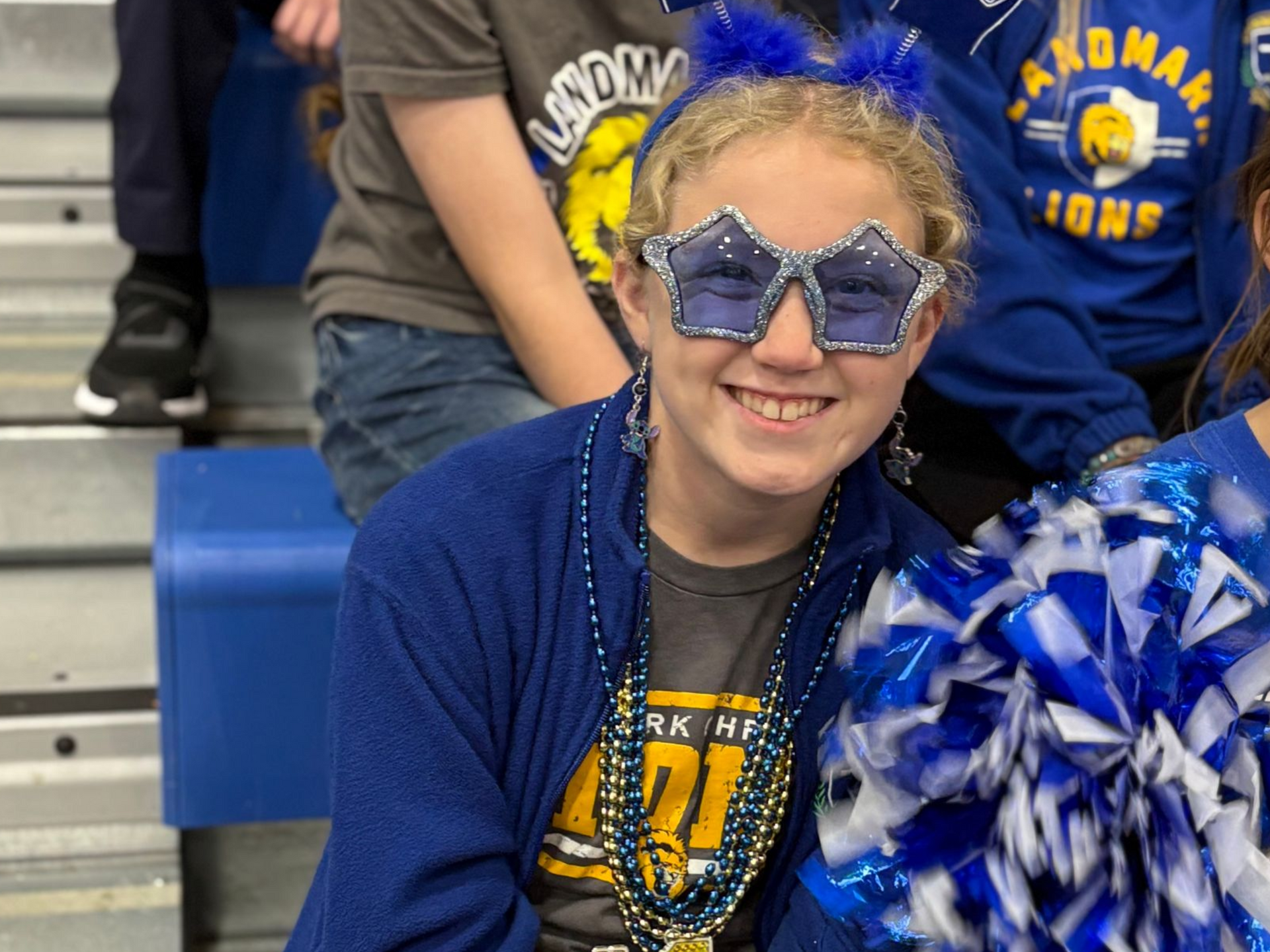 Smiling person in blue star sunglasses and pom-pom hat at a school event, holding blue-and-white pom-poms