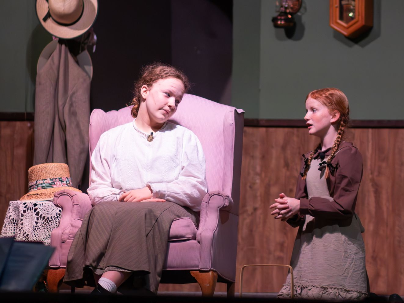 Two actors in period costumes onstage, one seated in a pink chair and one standing beside her.
