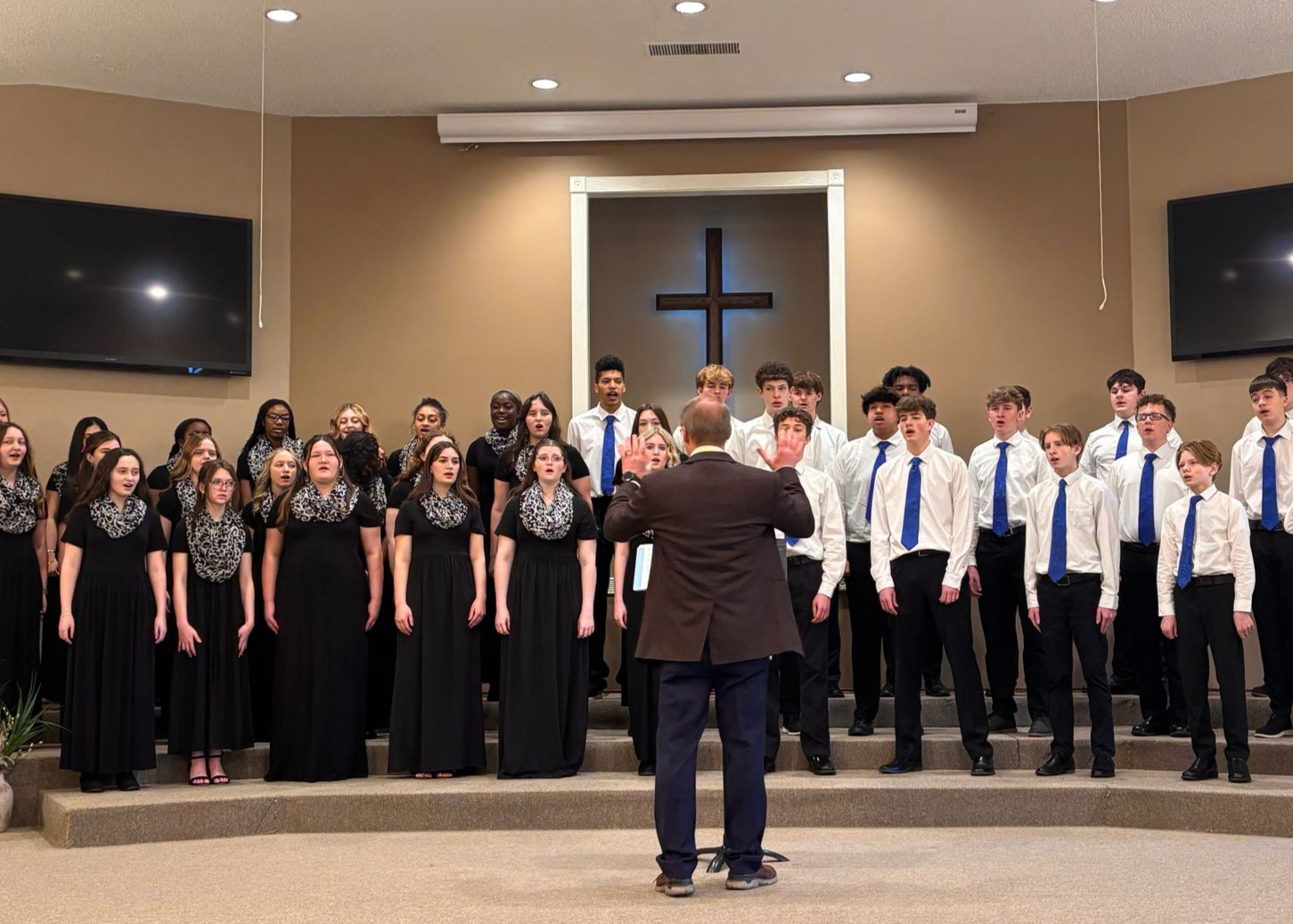 Choir performing on a church stage, conductor facing them with a cross in the background.