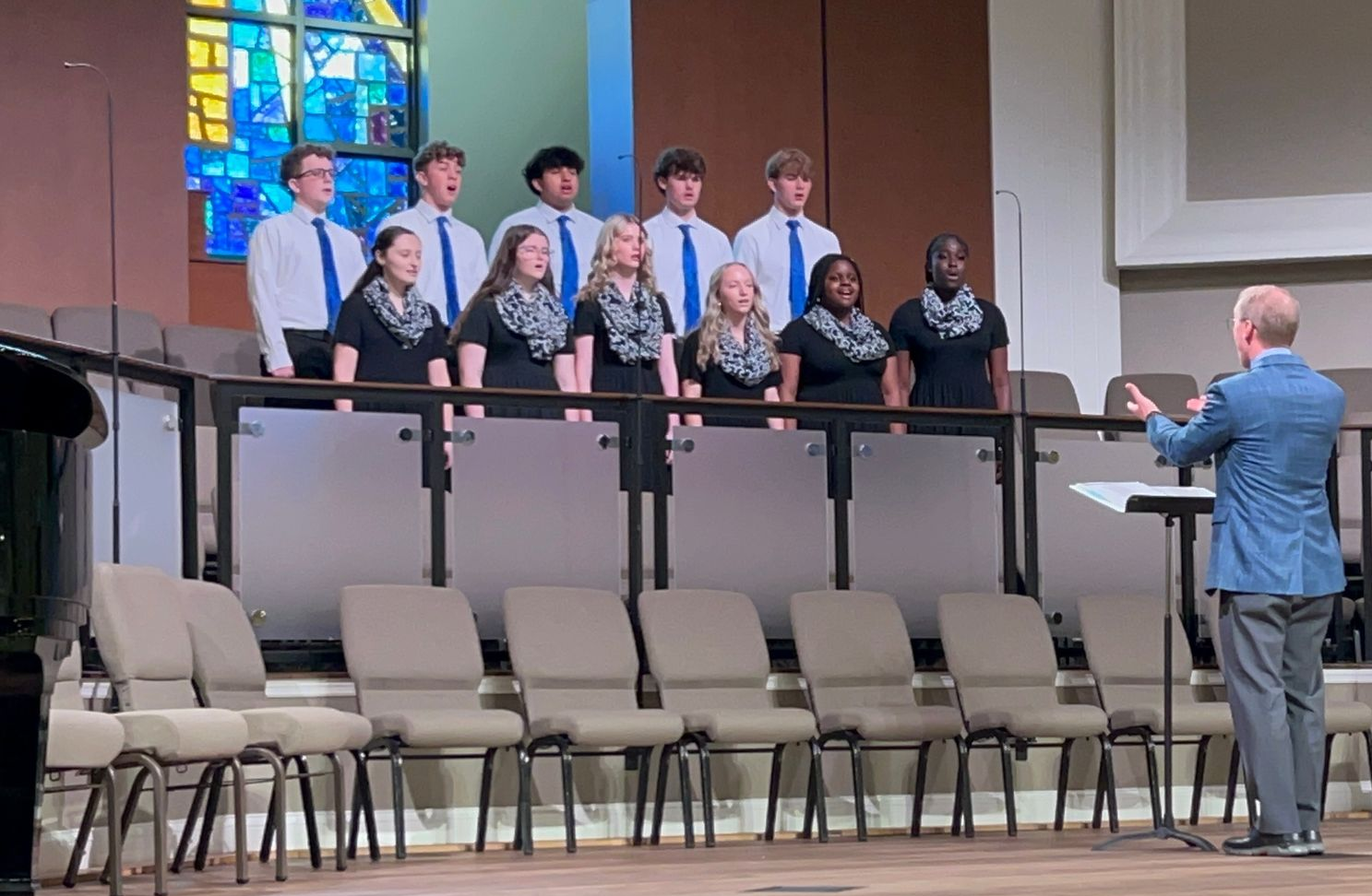 Choir on a church stage singing, with a conductor leading in front of empty chairs