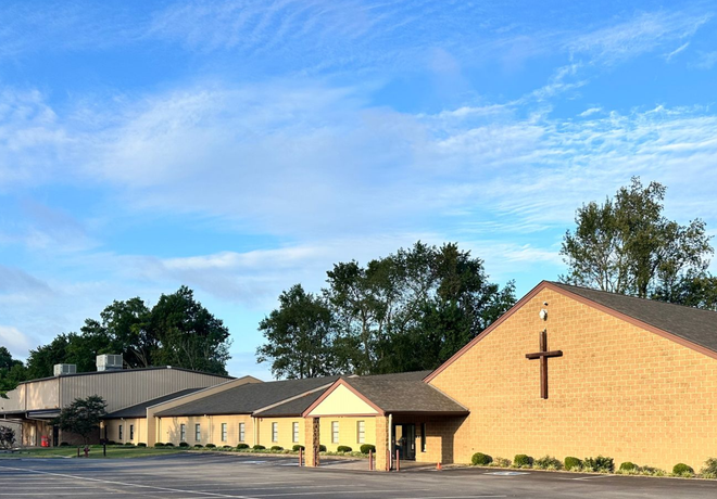 Church building with a cross on the front, set against a blue sky and trees.
