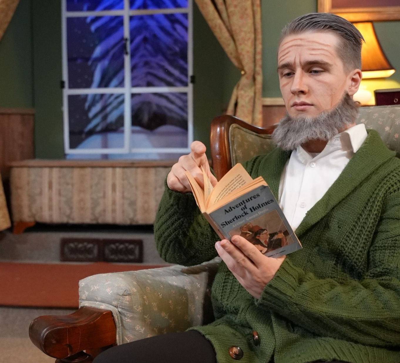 Man sitting in a chair reading a book in a cozy room by a window, wearing a green sweater and white shirt