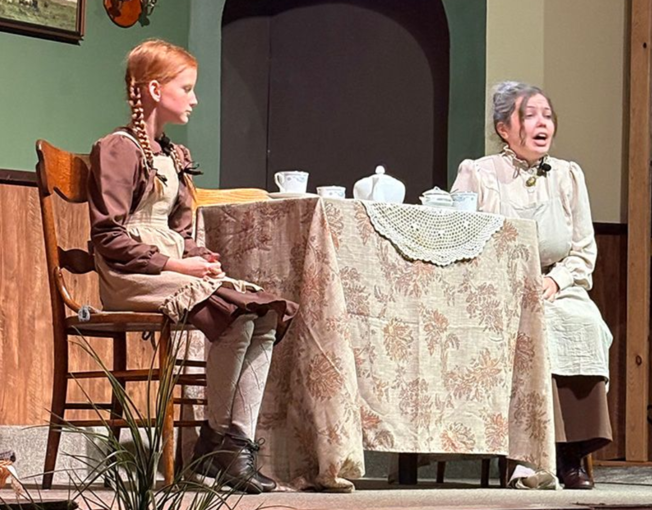 Two women seated at a small table on a stage, one speaking while the other listens.