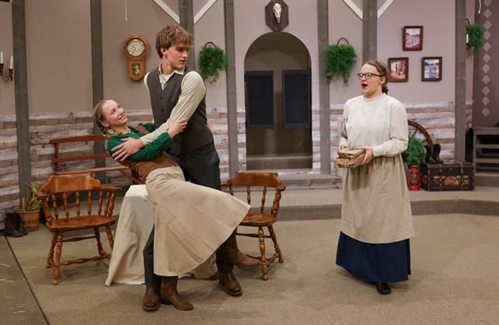 Man in green sweater hugs a woman twirling in a beige skirt; another woman stands nearby in a cozy room.