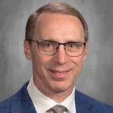 Portrait of a man in glasses and a blue suit with a tie, smiling against a gray background