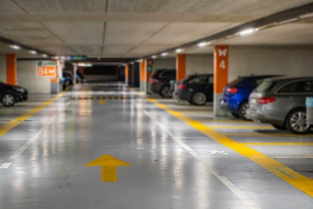 Estacionamento coberto com carros estacionados em ambos os lados e faixas demarcadas em amarelo no piso.