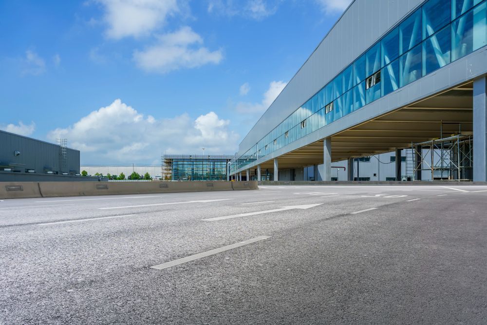 Terminal aeroportuário vazio, ao lado da calçada, sob um céu azul, com cobertura de vidro e ampla pista de asfalto.