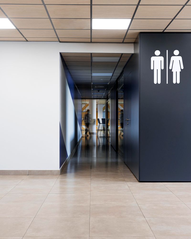Corredor moderno com placas de banheiro em uma parede escura e um corredor espelhado ao fundo.