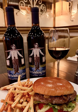 Two wine bottles, a glass of red wine, a burger, and fries on a table. The wine bottles' label depicts a person carrying buckets.