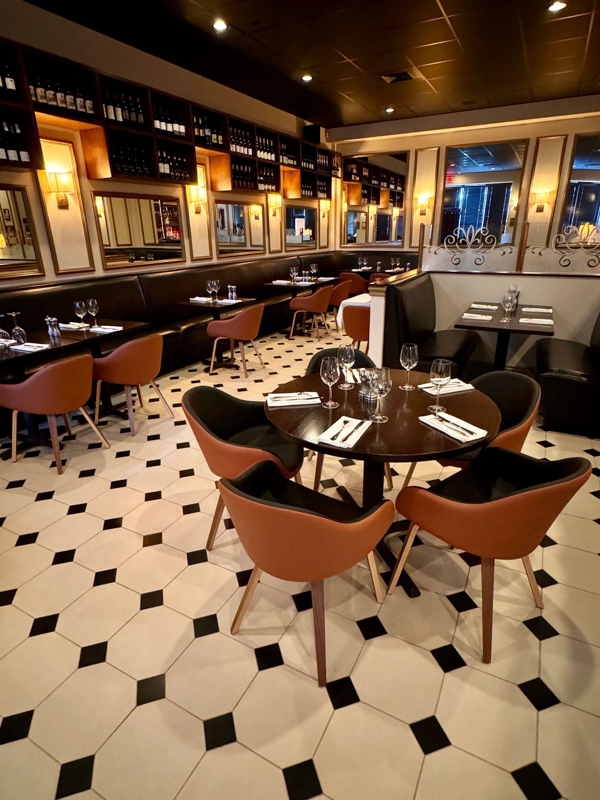 Restaurant interior with brown chairs around tables, black and white tiled floor, dark ceiling, mirrors.