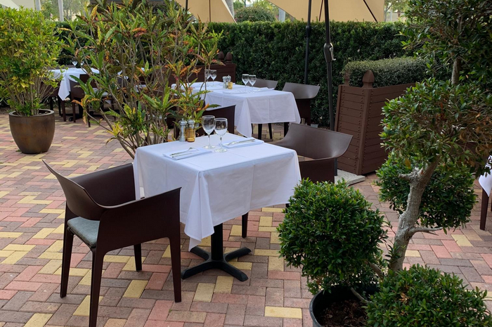 Outdoor restaurant tables with white tablecloths and chairs, surrounded by plants.