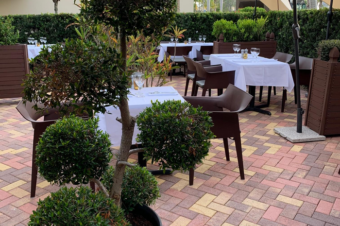 Outdoor dining area with tables set with white tablecloths, surrounded by trees and brick patio.