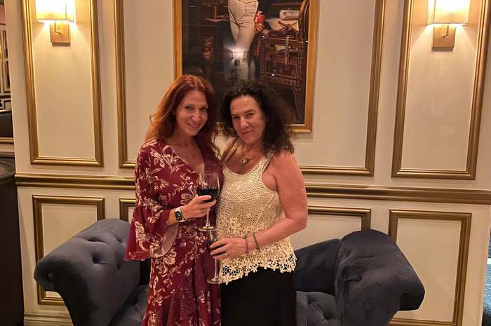 Two women pose by a velvet sofa, one holding wine, in a room with ornate wall paneling and a painting.