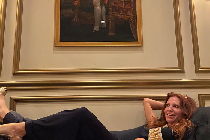 Woman reclining on a couch, looking to the side with a smile, under a framed painting on a gold-trimmed wall.
