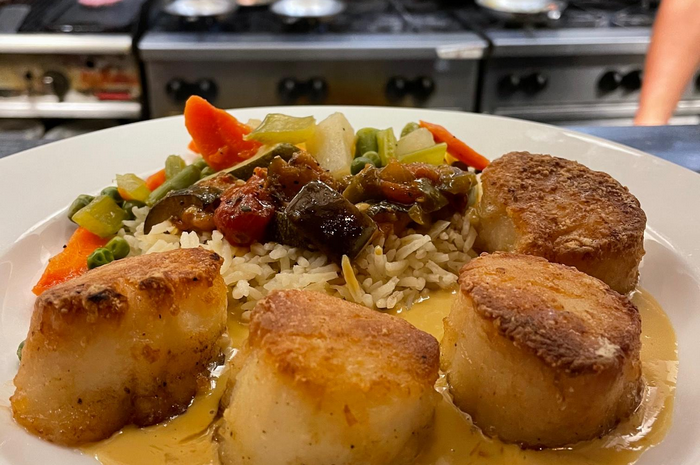 Seared scallops with rice and vegetables, on a white plate in a restaurant kitchen.