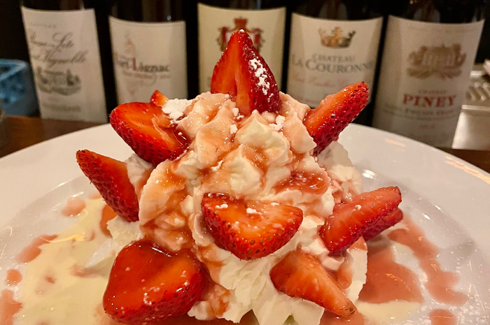 Strawberry shortcake on a white plate, topped with whipped cream and a strawberry sauce, with wine bottles in the background.