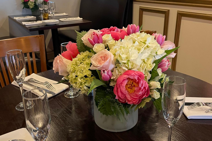 Floral arrangement on restaurant table; pink peonies, tulips, hydrangea, champagne glasses, setting.