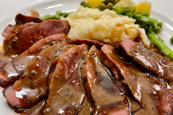 Sliced steak with gravy, mashed potatoes, peas, asparagus, and potatoes on a plate.