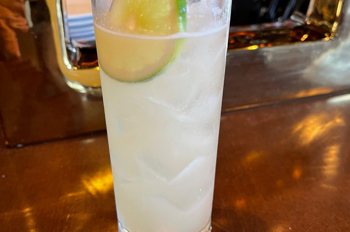 Tall glass of a light-colored cocktail with ice, garnished with a lime slice, sitting on a copper bar.