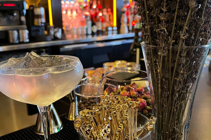 Cocktail on bar, ice, flowers, gold cocktail sticks, bottles in background.