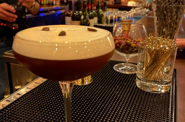 Espresso Martini in a stemmed glass, topped with foam and coffee beans, on a bar.