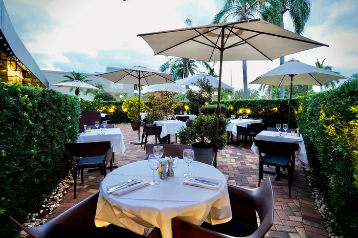 Outdoor restaurant patio with tables, white tablecloths, umbrellas, surrounded by hedges.