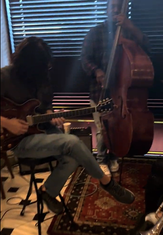 Two musicians playing instruments indoors: a guitarist seated and a bassist standing.