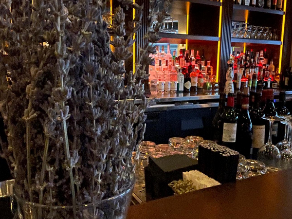 Dried lavender bouquet on a bar counter with liquor bottles and a brightly lit bar in the background.
