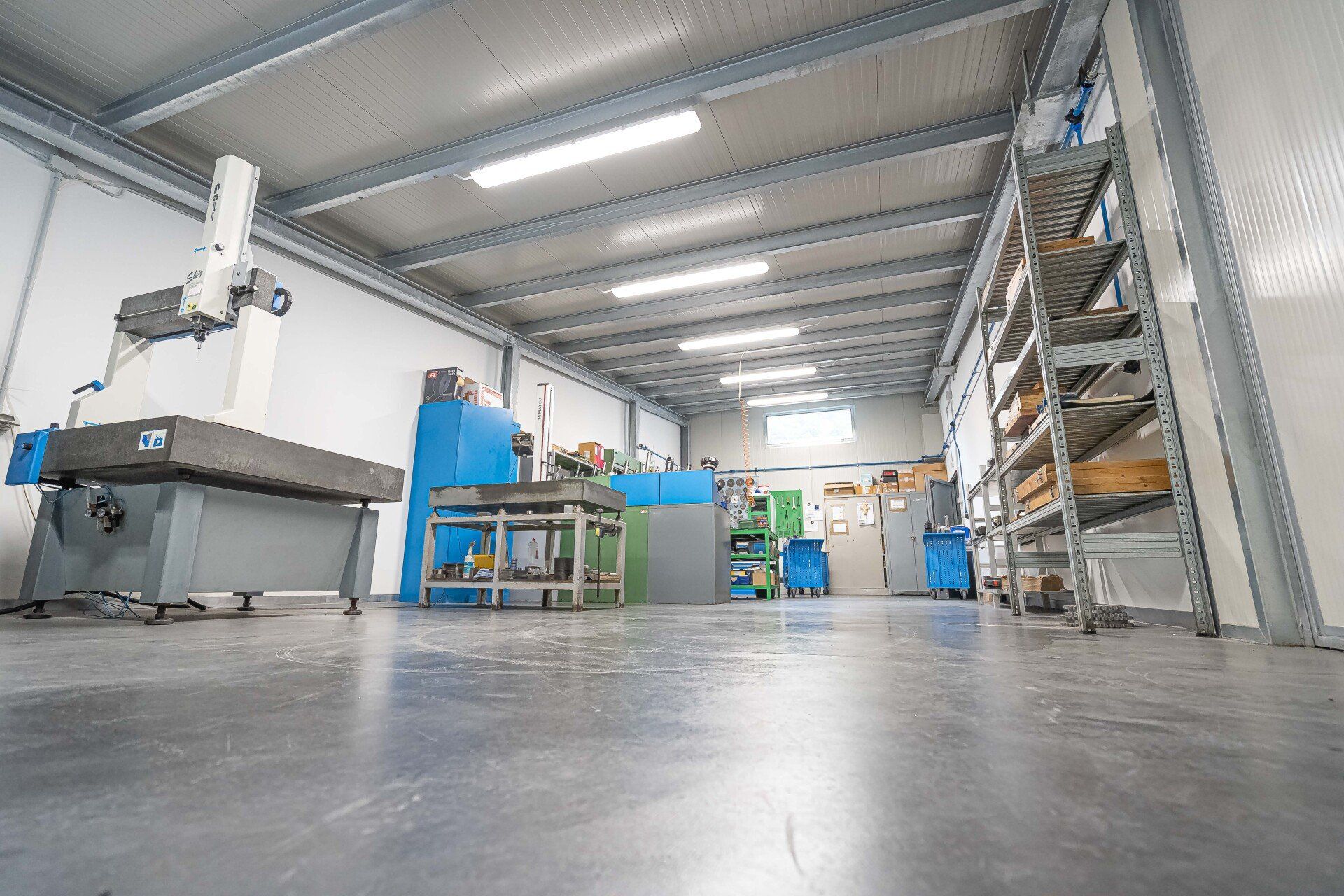 Interno di un'officina industriale con macchinari, banco da lavoro e scaffalature metalliche sotto luminose luci dall'alto.