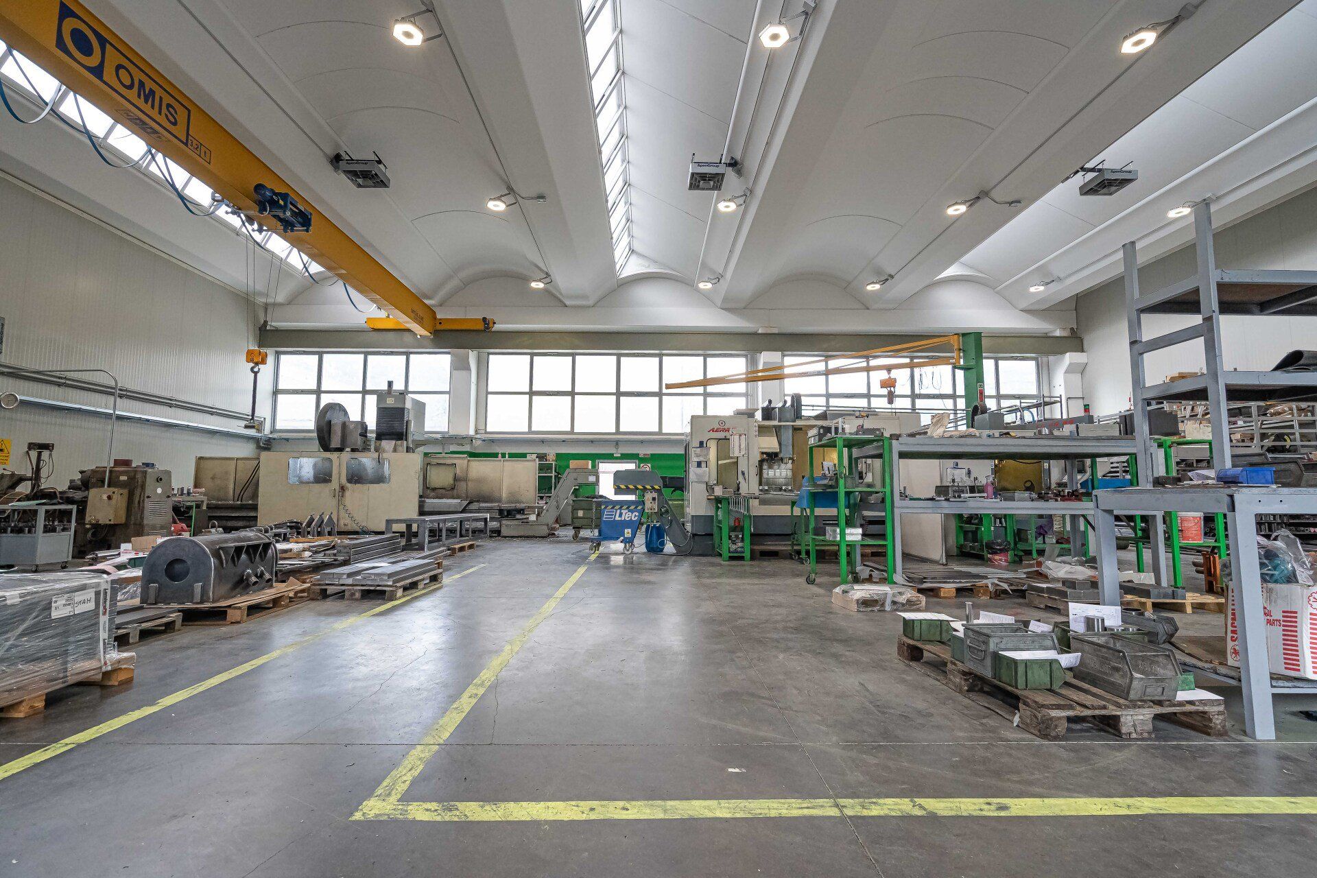 Ampio spazio industriale con macchinari, banchi da lavoro e illuminazione a soffitto; linee di sicurezza gialle delimitano il pavimento in cemento.