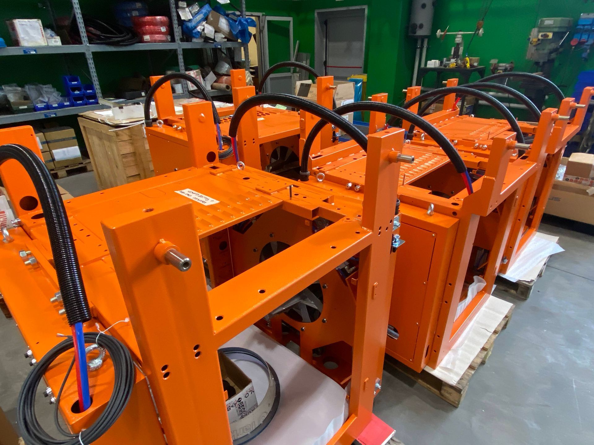 Telai di macchinari industriali arancioni su pallet in un'officina, con tubi e cavi collegati.