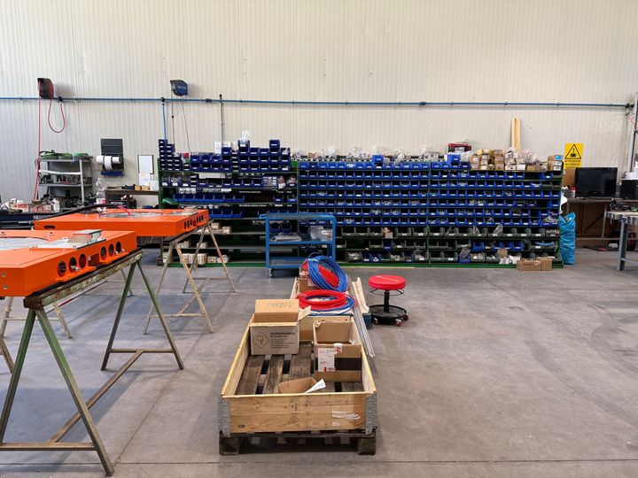 Officina con scafi di barche arancioni, scaffalature blu e un pallet di attrezzi su un pavimento di cemento.