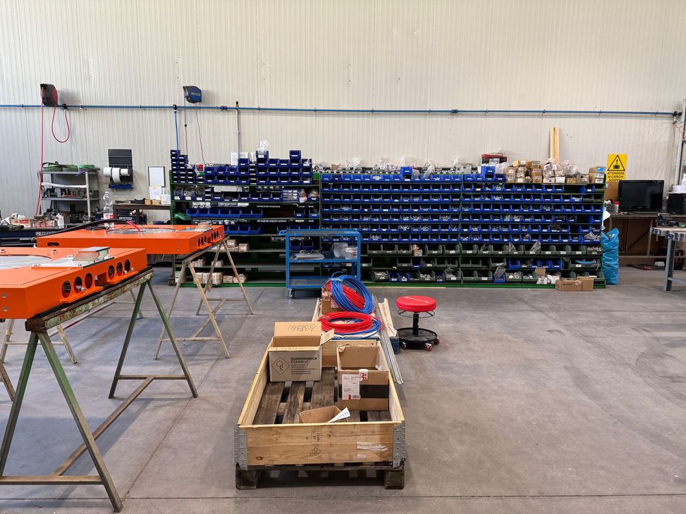 Officina con scafi di barche arancioni, scaffalature blu e un pallet di attrezzi su un pavimento di cemento.