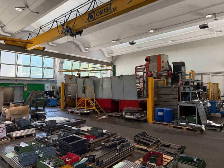 Officina industriale con attrezzi e attrezzature sparsi, gru a ponte gialla e scaffalature metalliche lungo la parete.