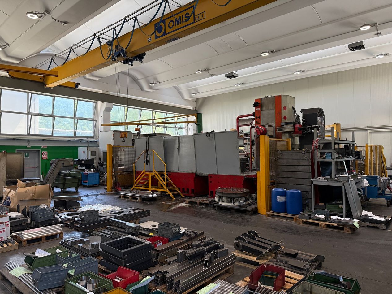 Officina industriale con attrezzi e attrezzature sparsi, gru a ponte gialla e scaffalature metalliche lungo la parete.