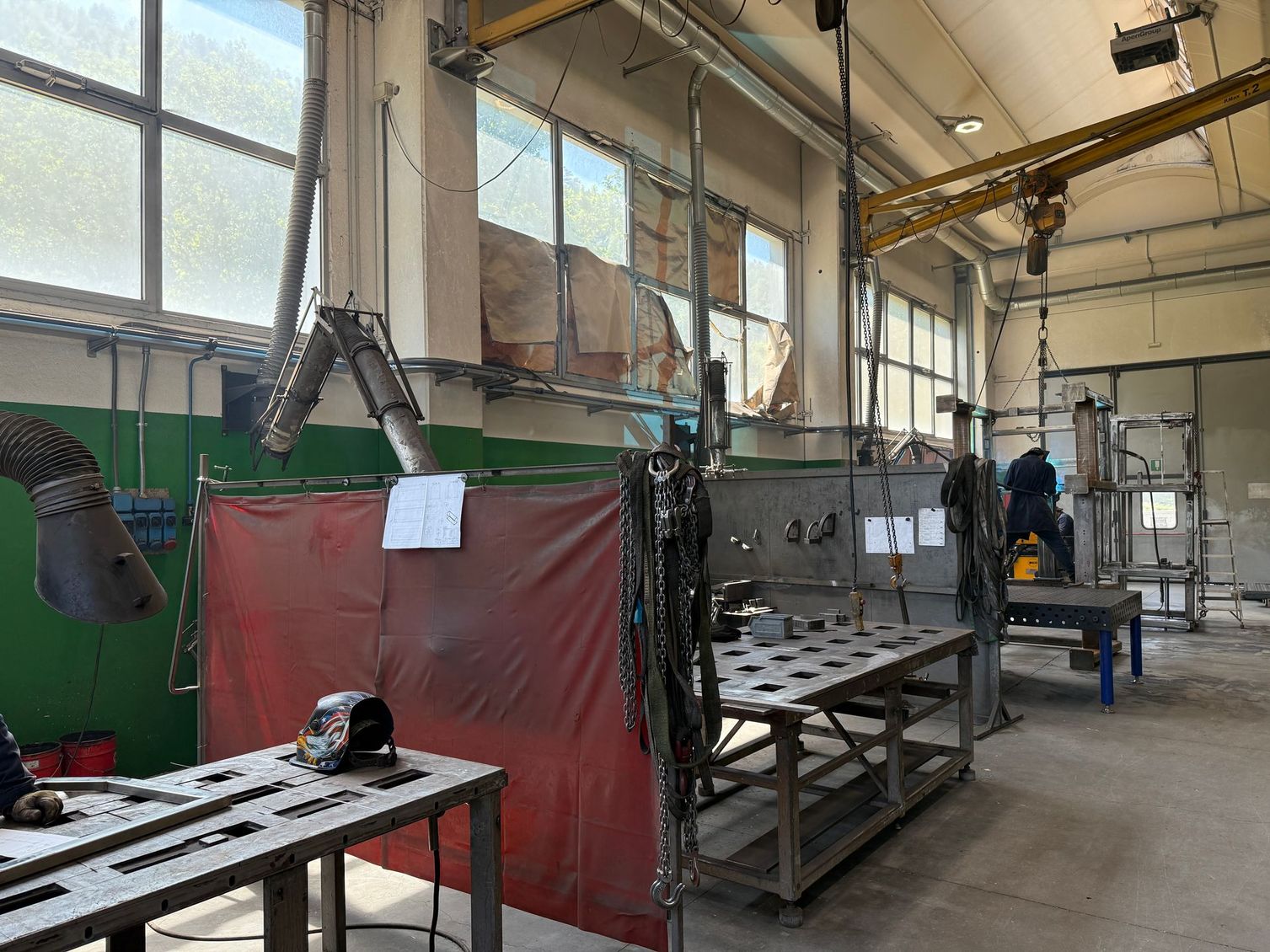 Officina industriale con tavoli metallici, tenda da saldatura rossa, condotti sospesi e un operaio vicino ai macchinari.