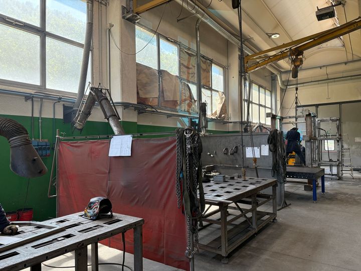 Officina industriale con tavoli metallici, tenda da saldatura rossa, condotti sospesi e un operaio vicino ai macchinari.