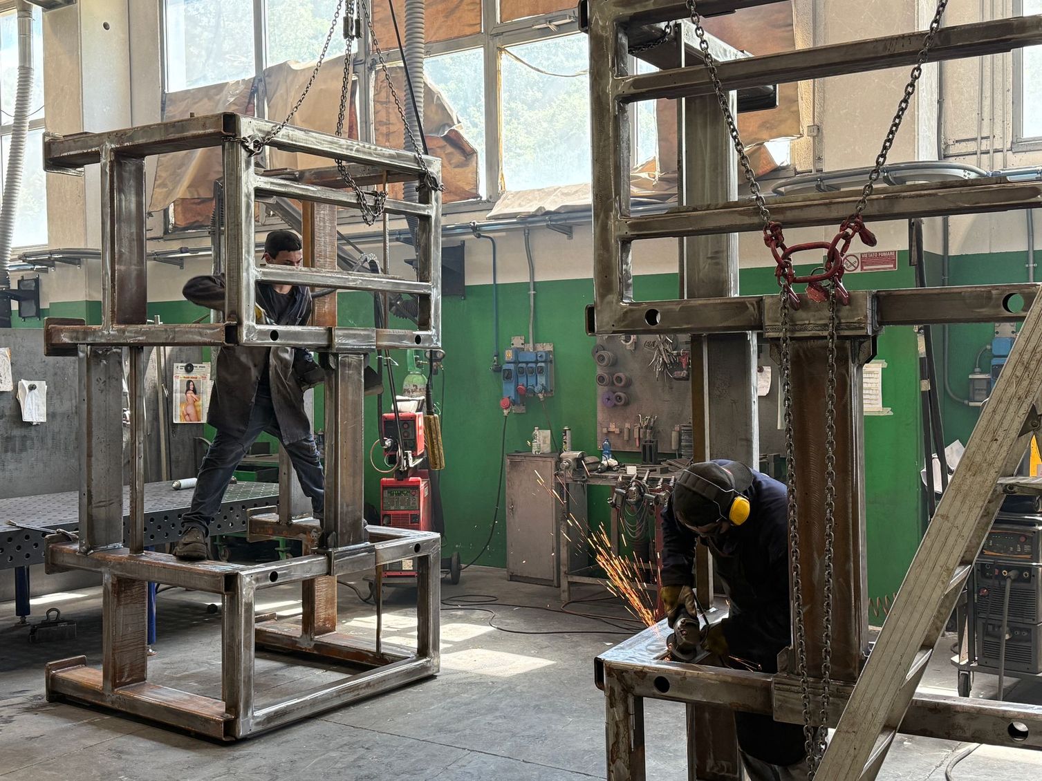 In un'officina, alcuni operai saldano grandi telai metallici, con scintille che volano vicino a una scala e a un'impalcatura.