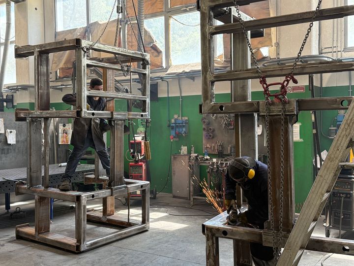 In un'officina, alcuni operai saldano grandi telai metallici, con scintille che volano vicino a una scala e a un'impalcatura.