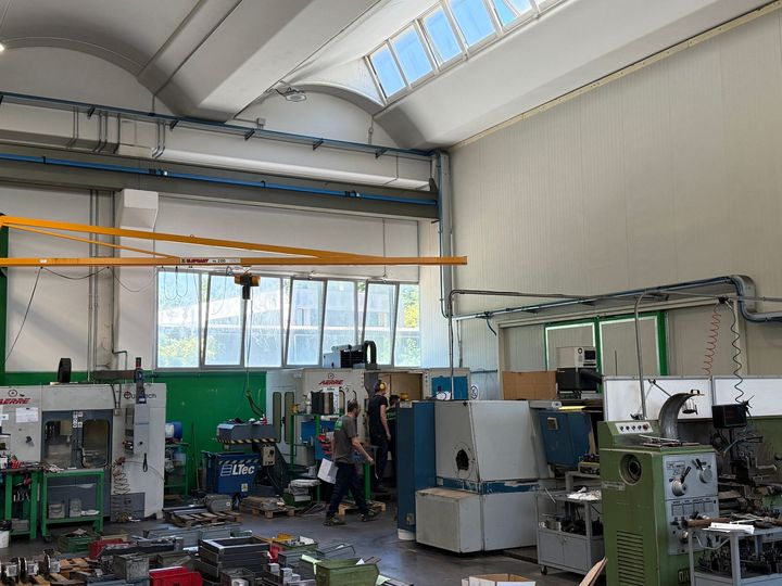 Officina industriale con macchinari, operai e grandi finestre sotto un soffitto alto illuminato da un lucernario.