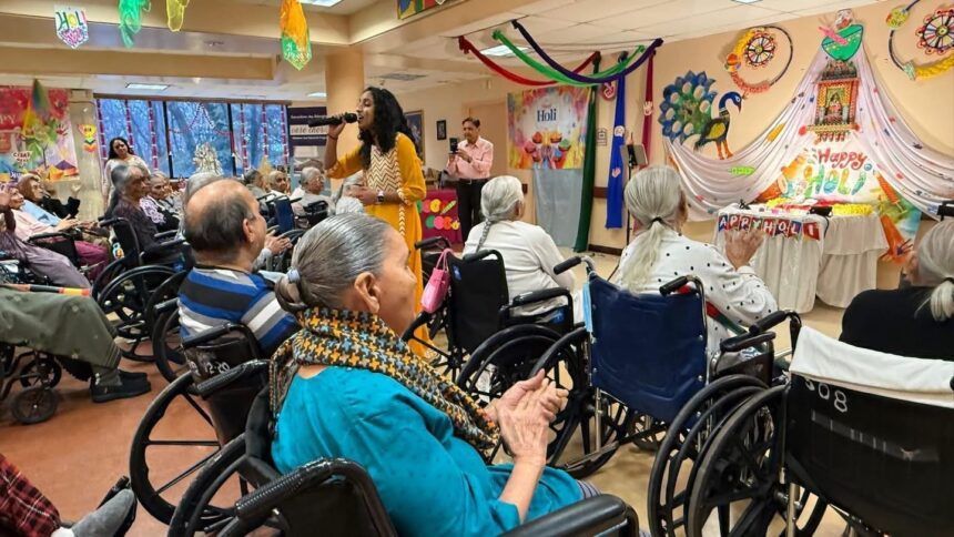 A performer sings into a microphone for an audience of seniors in wheelchairs gathered in a decorated community room.