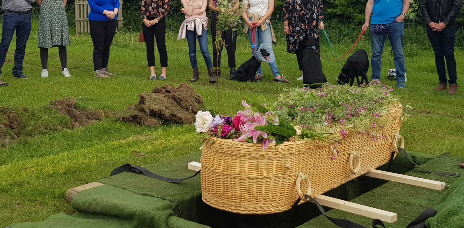 A group of people are standing around a coffin in the grass.