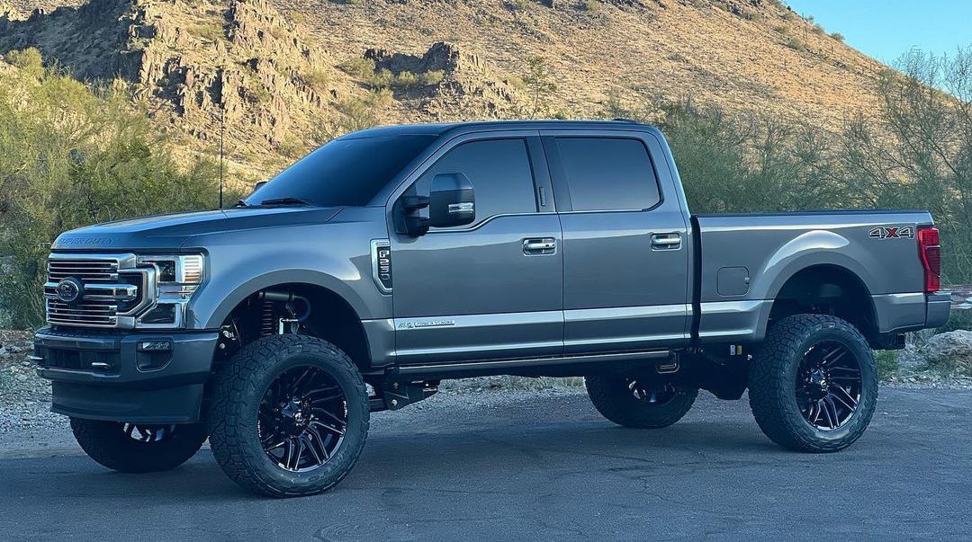 Gray lifted Ford pickup truck with black wheels, parked in front of a mountain.