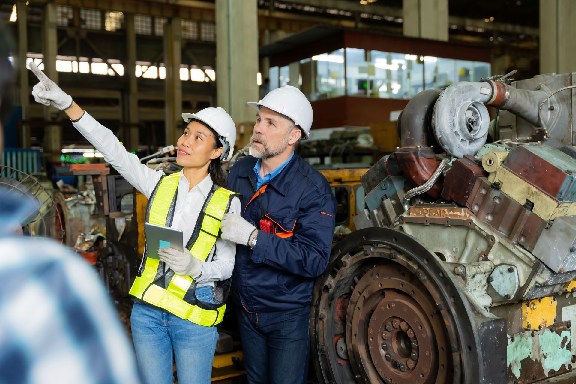 Woman in vest and hard hat points, discussing with a man in a factory setting.