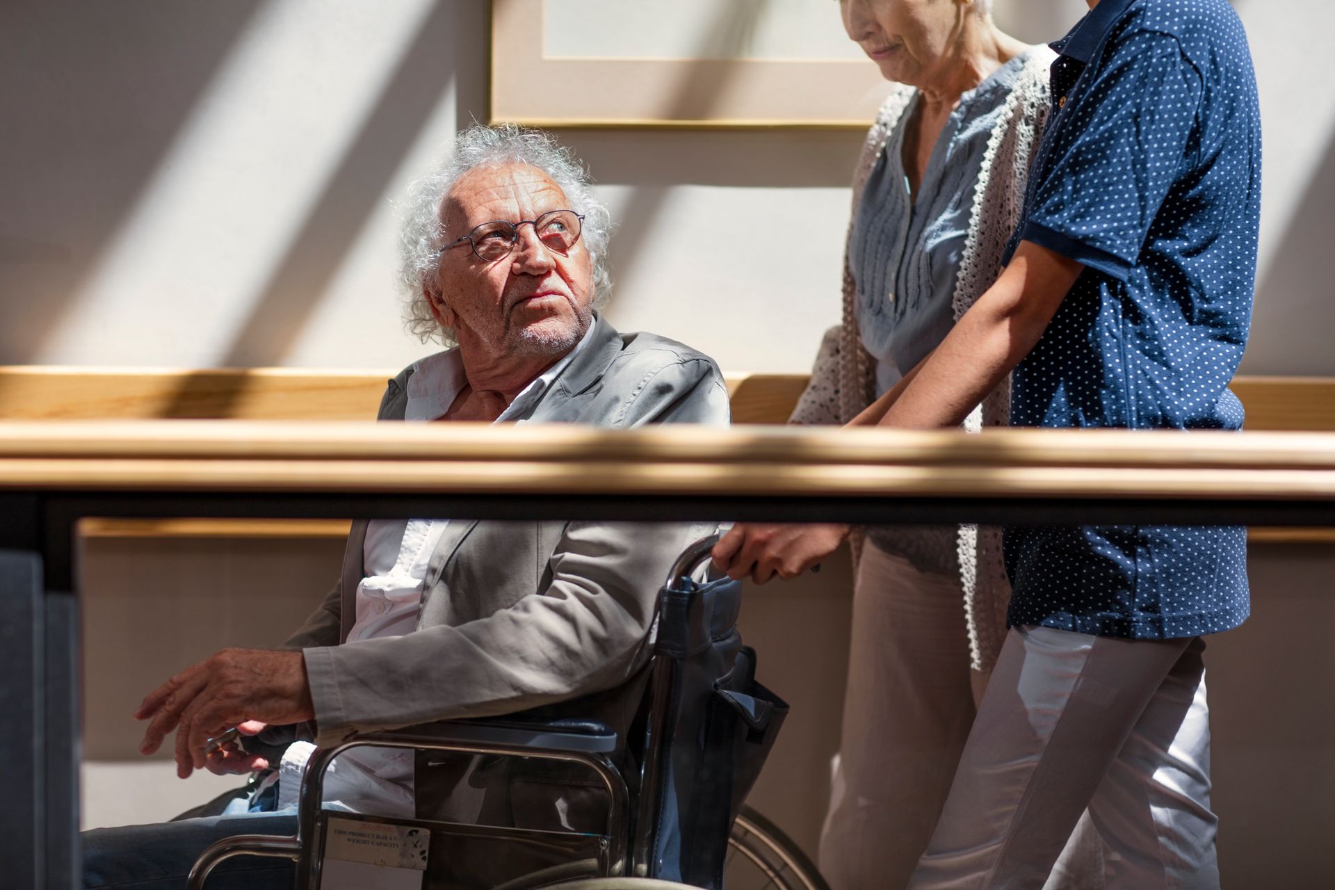 Man in wheelchair with two people, indoors near a wooden railing.