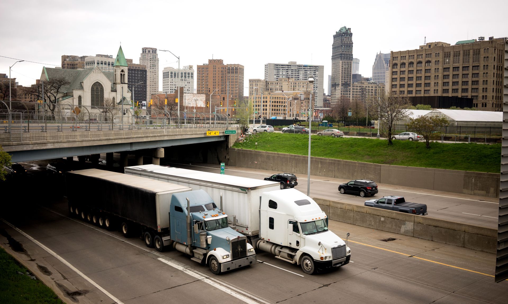 Two semi-trucks on a highway under an overpass in a city, with buildings in the background.