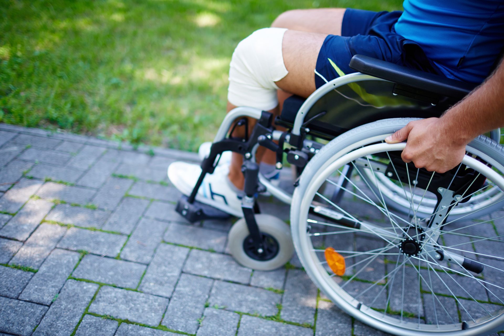 Person in wheelchair with bandaged knee on a brick path, holding the wheel.